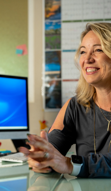 A middle-aged woman with long blond hair sits at a desk and smiles.