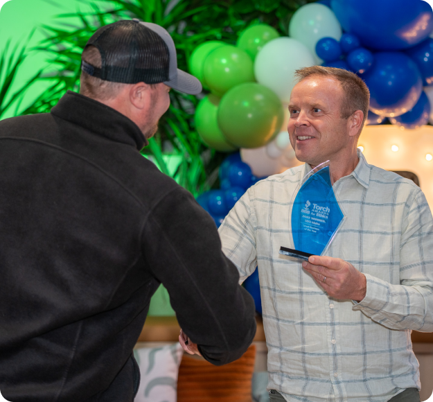A young man holds a blue glass award while shaking hands with the man who presented it to him.