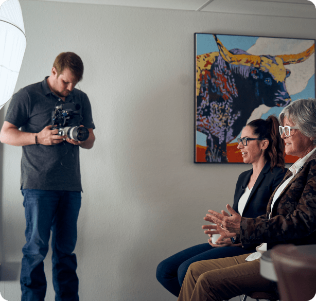 A younger and an older woman sit next to each other and smile as a photographer takes their photograph.