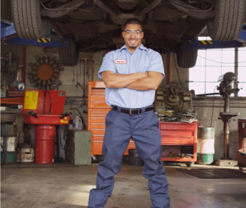A smiling young mechanic stands with his arms folded in a car repair garage.