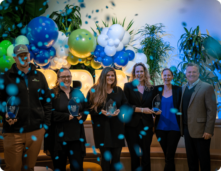 A group of men and women stand together holding blue glass awards, surrounded by balloons and confetti.