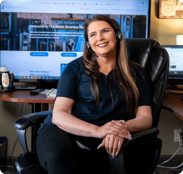 A young woman wearing a microphone headset smiles as she sits in a chair with a large computer screen in the background.