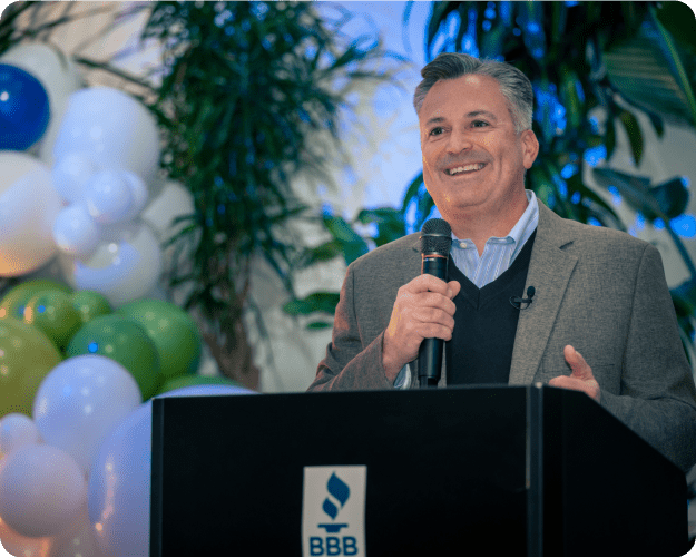 A man stands at a Better Business Bureau logoed podium with a microphone in his hand and balloons in the background.
