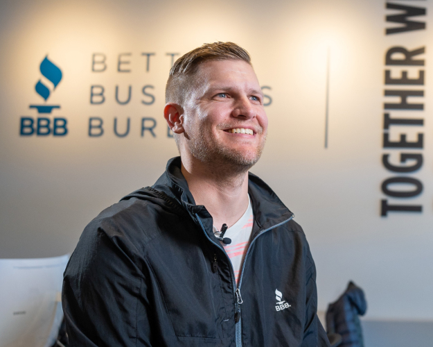 A male Better Business Bureau employee sits in an office with the company’s mission statement on a wall in the background.