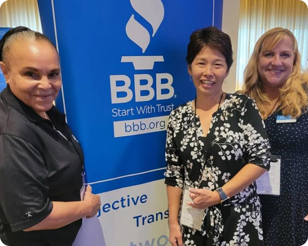 Three female Better Business Bureau teammates stand together, smiling, next to a Better Business Bureau pull-up banner.